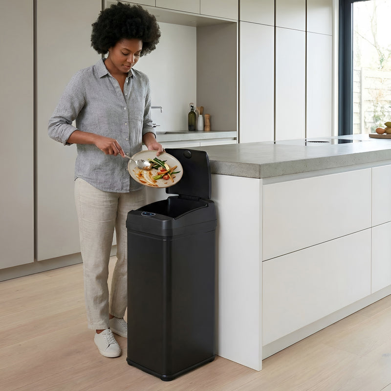 A woman in a modern kitchen using the iTouchless DZT13PBK black stainless steel 50L sensor trash can to throw garbage away.