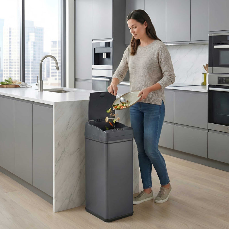 A woman in a modern kitchen using the iTouchless DZT13PGY gray stainless steel 50L sensor trash can to throw garbage away.