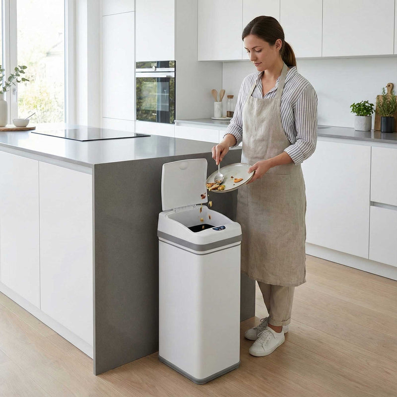 A woman in a modern kitchen using the iTouchless DZT13PWH white stainless steel 50L sensor trash can to throw garbage away.