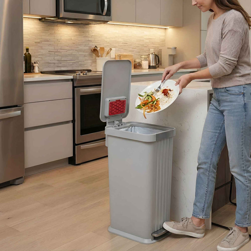 A woman in a modern kitchen using the iTouchless PE13OO stone gray step pedal trash can 50L to scrape food scraps off a plate.