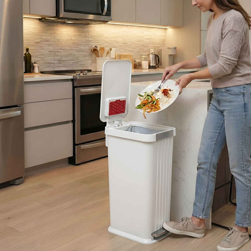 A woman in a modern kitchen using the iTouchless PE13WW chalk white step pedal trash can 50L to scrape food scraps off a plate.