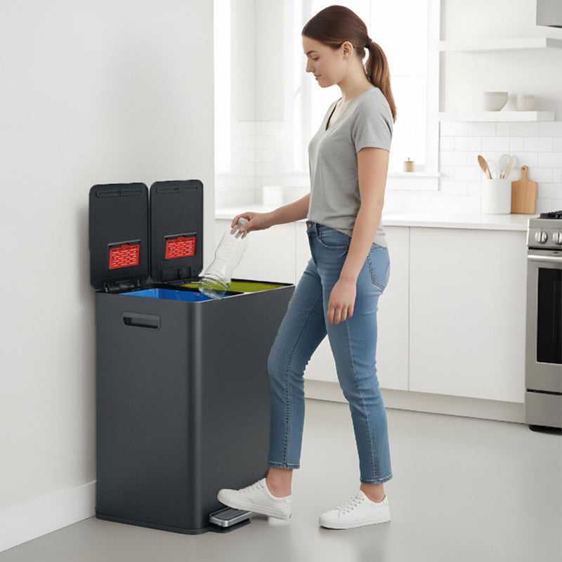 A woman in a modern kitchen using the iTouchless PC60RSPGY gray stainless steel 60L step trash can to sort a plastic bottle into one of the recycling compartments.