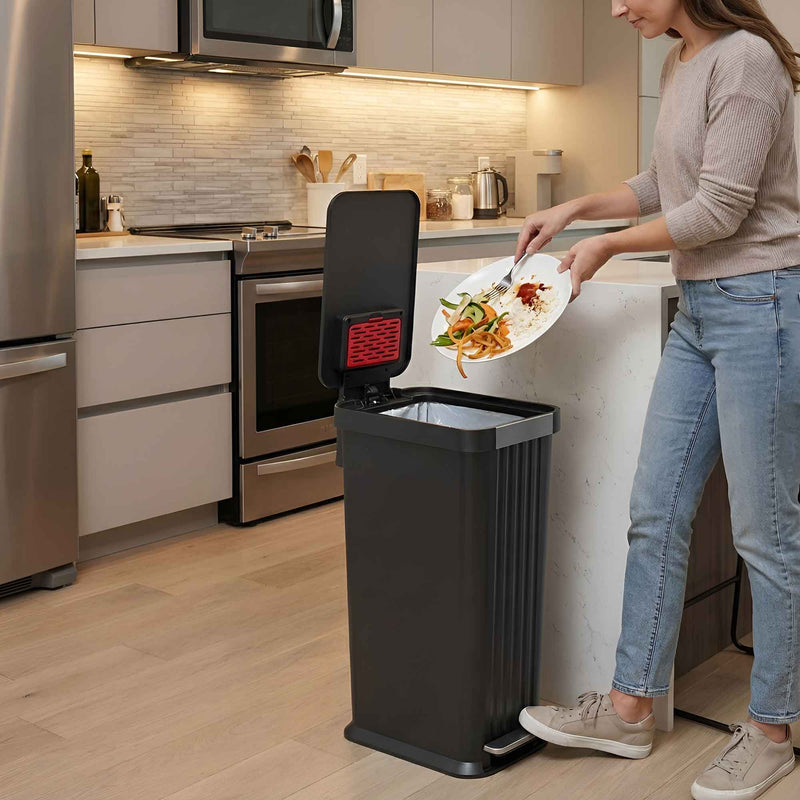 A woman in a modern kitchen using the iTouchless PE13BB black step pedal trash can 50L to scrape food scraps off a plate.